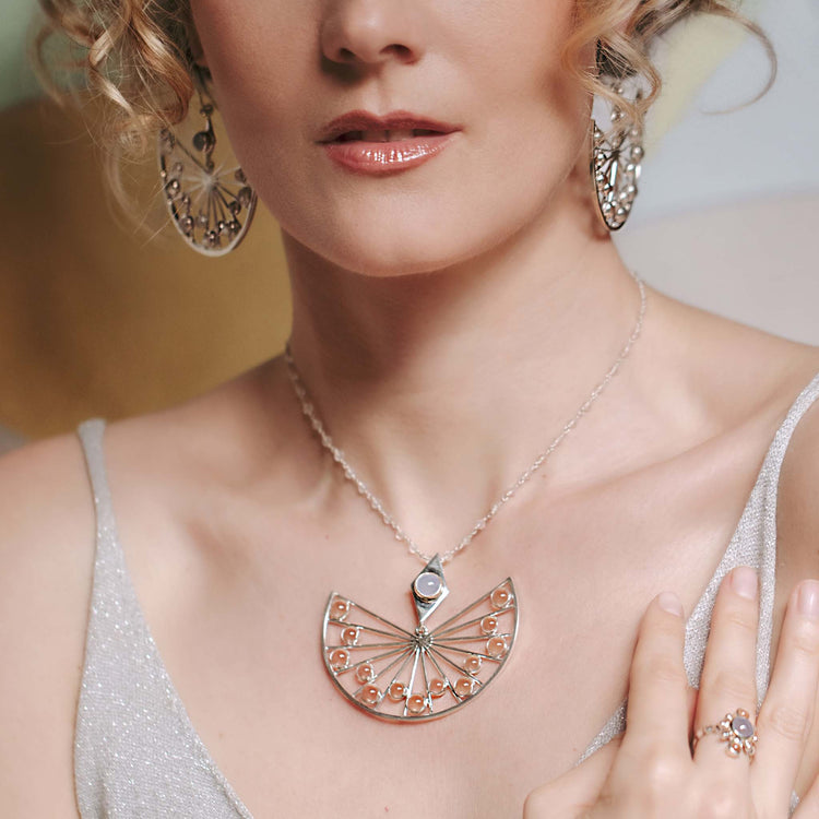 A woman wearing a glittery silver dress is adorned with an intricately designed Kaora Sandara Jewelry Higher Self Pendant - Sterling Silver with Crystal & Chalcedony, featuring a pendant with multiple spokes and circular elements. She also wears large circular earrings and a ring with a floral design. Her blonde hair is styled in loose curls.