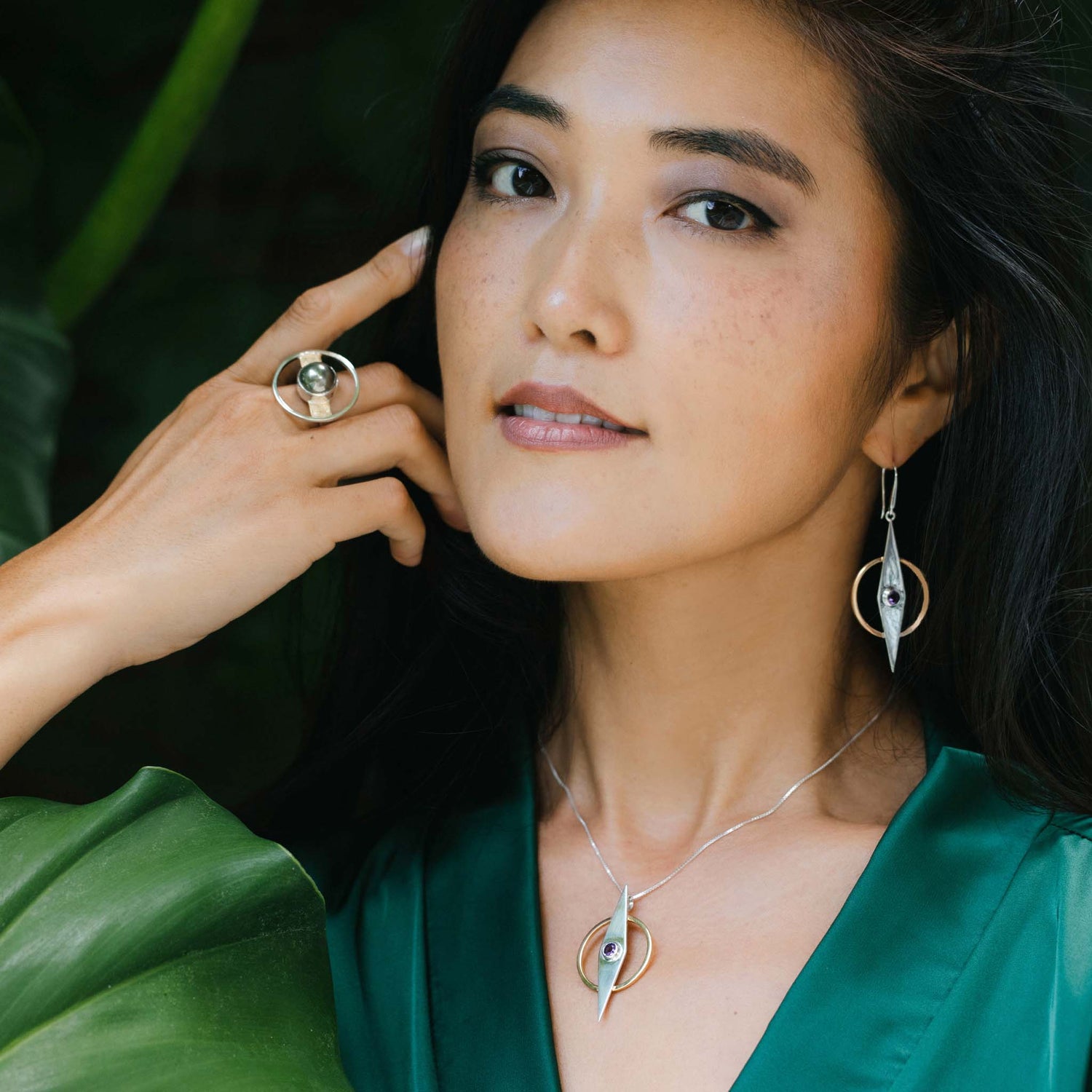 A woman with long dark hair is wearing a green blouse and matching Sterling Silver 925 jewelry: a ring, earrings, and the Intuition Pendant – Sterling Silver & Gold-Plated with Amethyst by Kaora Sandara Jewelry. The jewelry features a modern design with circular and teardrop shapes. She poses with her hand near her face, showcasing the Intuition series ring and earrings against a leafy background.