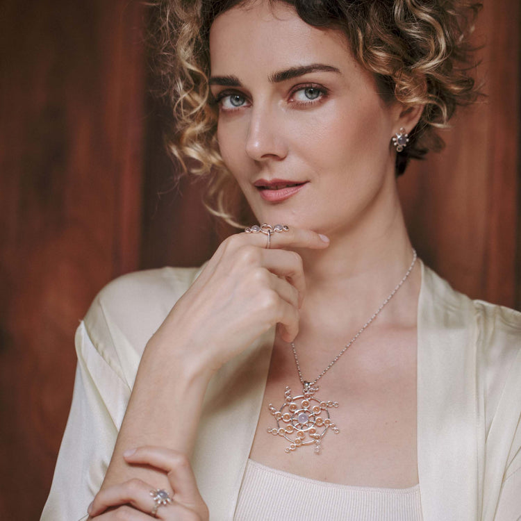 A woman with curly hair, dressed in a cream blouse and adorned with intricate jewelry from Kaora Sandara Jewelry, including the Perfection of Life Pendant - Sterling Silver with Crystal & Chalcedony, along with matching earrings and rings, poses with a slight smile against a wooden background. Her hand gently touches her chin.