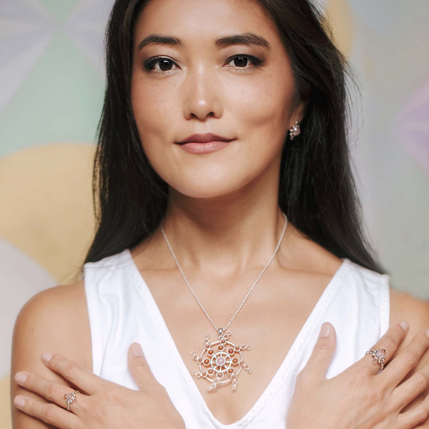 A woman with long dark hair is wearing a white sleeveless top. She has a calm expression and wears the Perfection of Life Pendant from Kaora Sandara Jewelry, an intricate piece of sterling silver adorned with crystal and rose quartz. She also sports matching rings. Her arms are crossed over her chest, showcasing the jewelry against a softly colored geometric background.