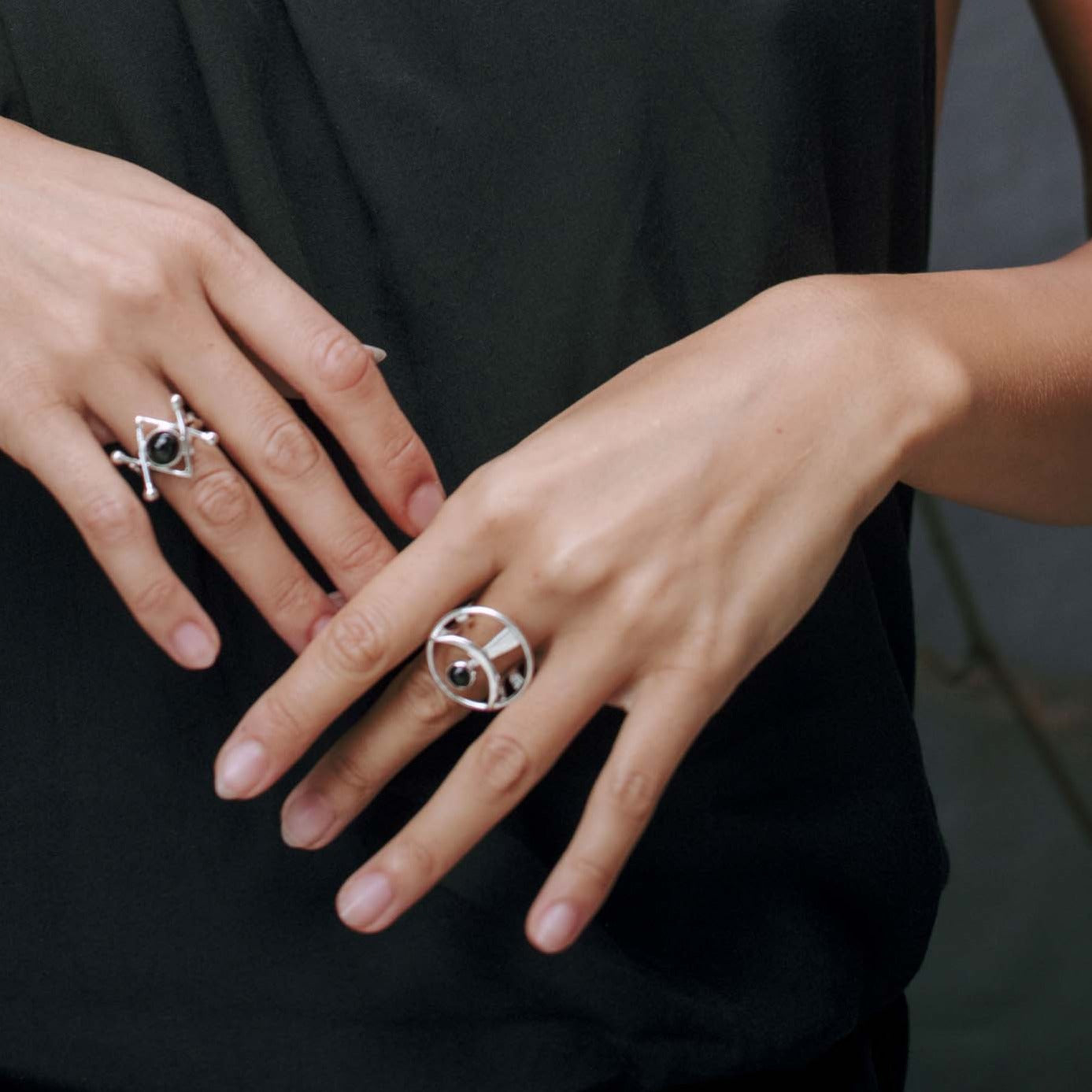 A close-up of a person's hands and wrists adorned with sterling silver jewelry. The left hand features a ring with a geometric, crisscross design, while the right hand boasts the "Releasing From the Deepest Fears Ring" in sterling silver with black agate from Kaora Sandara Jewelry. The person is wearing a sleeveless black top.