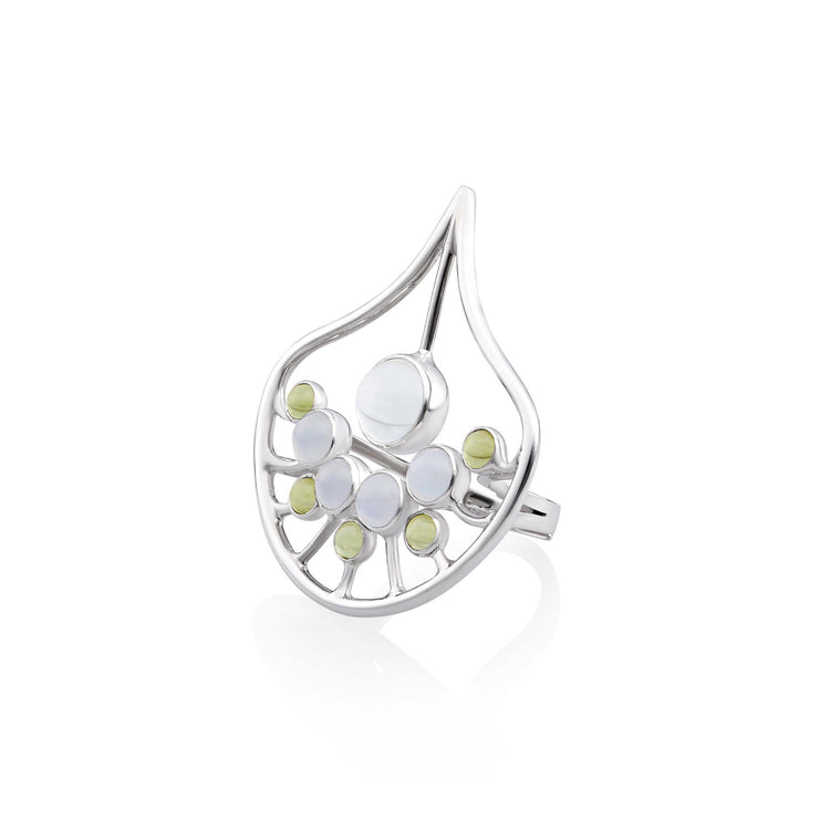 The Body Relaxation Ring by Kaora Sandara Jewelry is a sterling silver ring featuring an abstract design with a teardrop-shaped frame, perfect for enhancing energy flow. Inside the frame are several round crystal and chalcedony stones, with a larger central white stone and smaller green peridot and white stones arranged below it. The background is white.