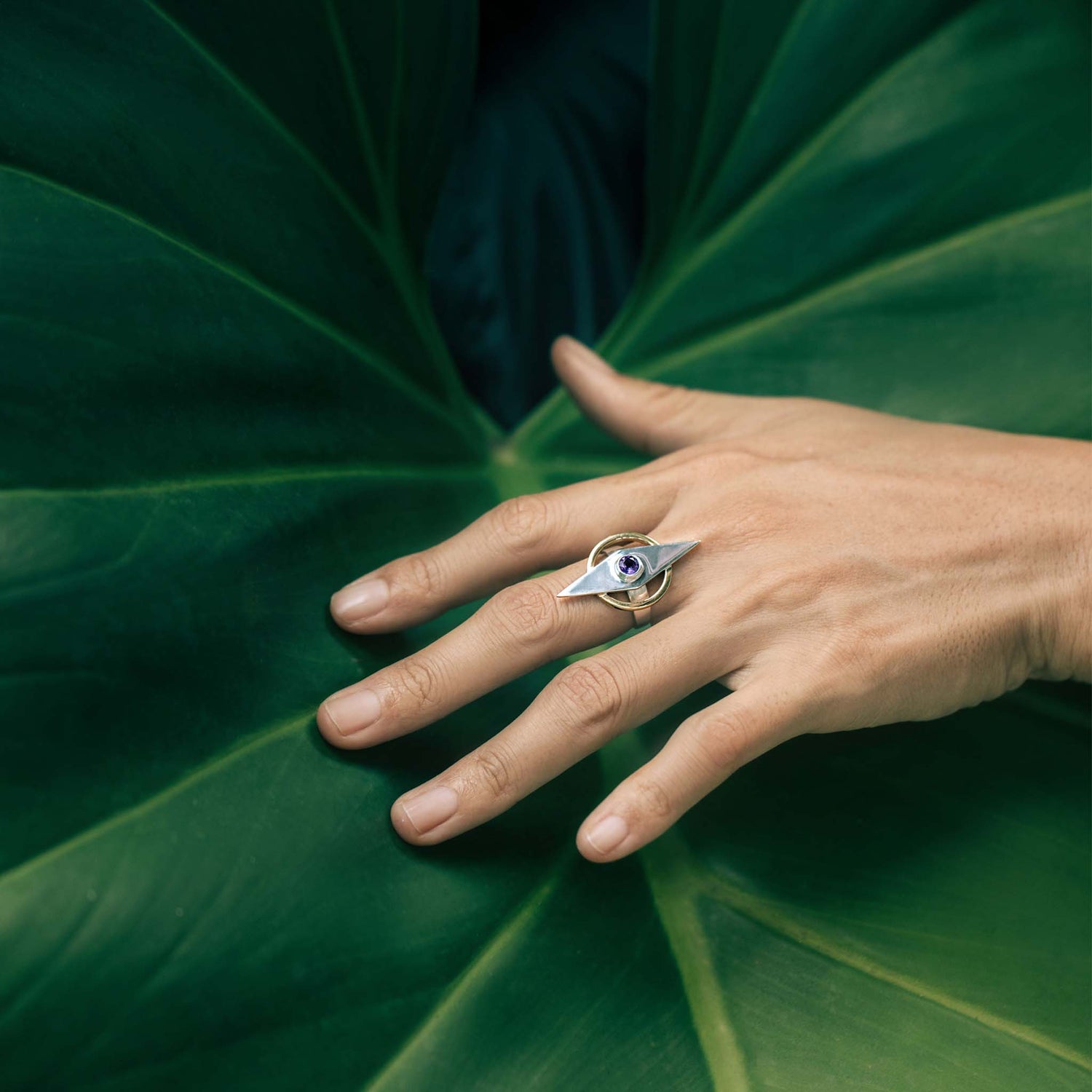 A hand adorned with the Intuition Ring from Kaora Sandara Jewelry, made of sterling silver and gold-plated with an amethyst gemstone, rests on a large green leaf, creating a striking contrast between the elegant jewelry and the lush, natural background.