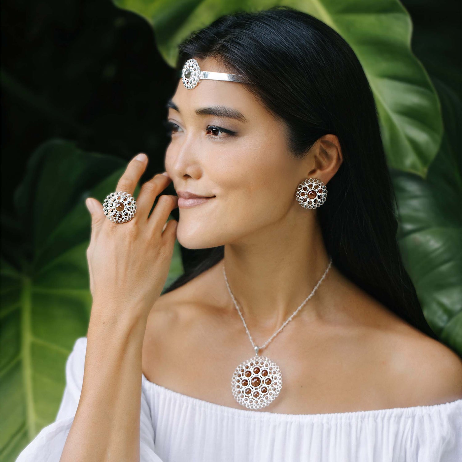 Profile close-up of a model in nature, set against a leafy background. The model is wearing a tiara, earrings, a pendant, and a ring from the Purity series in size L. These silver pieces are round in shape, featuring small round rock crystal stones and a single large central round rock crystal.