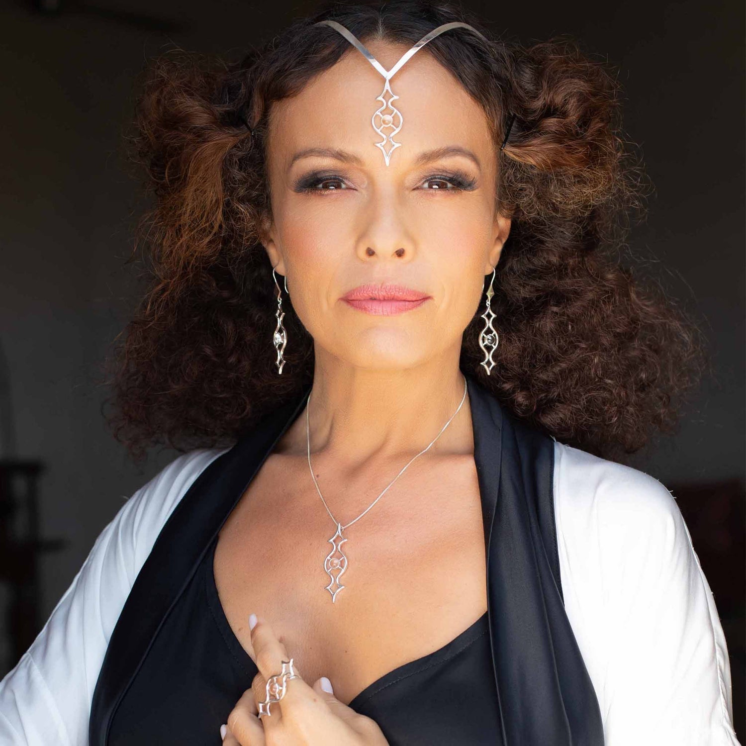 A person with curly hair, adorned in the intricate Quantum Shift NEW sterling silver jewelry with crystal gemstones by Kaora Sandara Jewelry, including a headpiece, earrings, necklace, and rings in size S, poses confidently. Dressed in stylish black and white attire against a dark background, the lighting accentuates their facial features and the sparkling rhinestone crystals of their accessories.