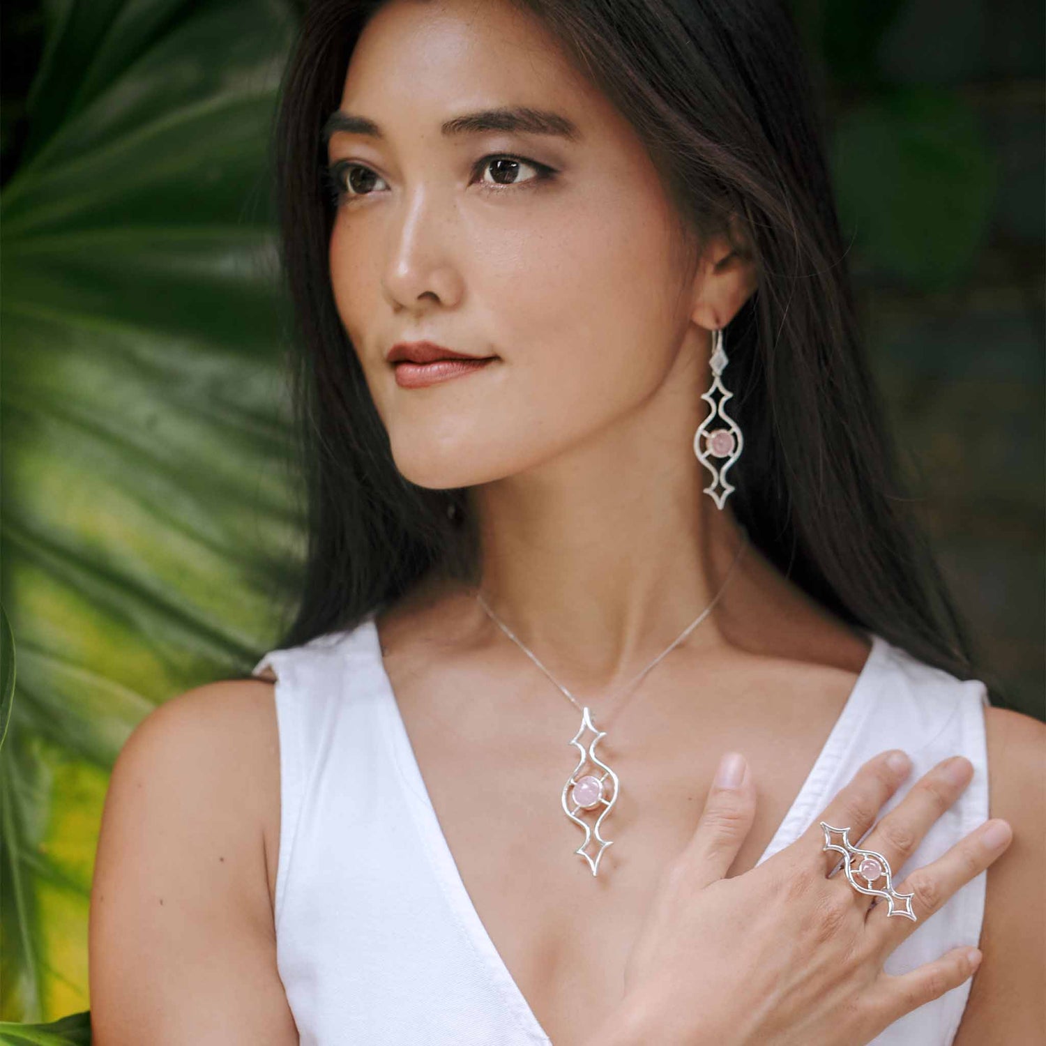 A woman with long, dark hair stands in front of lush greenery. She wears a white sleeveless top and Kaora Sandara Jewelry’s Quantum Shift NEW Earhooks, sterling silver 925 with rose quartz, along with a matching pendant necklace and ring. One hand is raised to her chest, showcasing the intricate design of the ring.