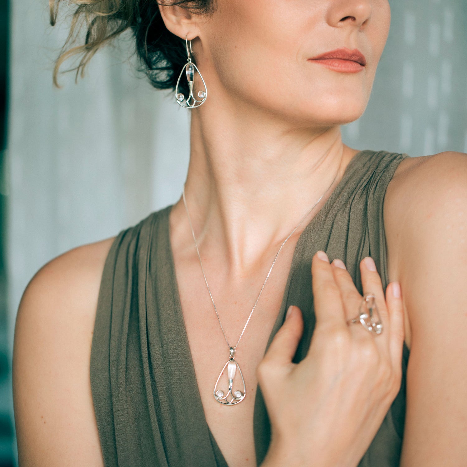 A woman wearing an elegant sleeveless green dress showcases a matching sterling silver jewelry set from Kaora Sandara Jewelry, including the Self-Worth Pendant - Sterling Silver with Crystal necklace, earrings, and a ring. She poses gracefully with one hand resting on her chest, highlighting the intricate designs.