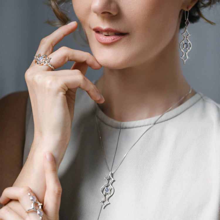 A woman models an elegant jewelry set from Kaora Sandara Jewelry, including a long pendant necklace, ornate drop earrings, and multiple rings featuring Sterling Silver 925. She gently touches her face and neck, showcasing the intricate designs of the accessories against her light-colored sleeveless top. Front and center are the Quantum Shift NEW Earhooks – Sterling Silver with Chalcedony in Size S.