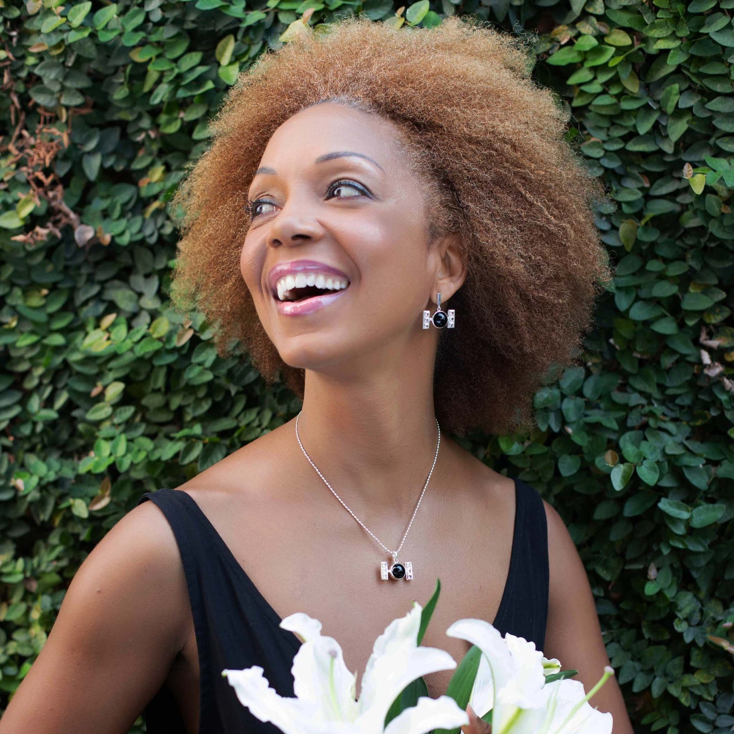 A woman with curly hair smiles joyfully while holding white flowers. She is wearing a black dress and matching jewelry from Kaora Sandara Jewelry, featuring the Silver Earhooks Health Formula – Black Agate & Crystal. The earrings are shaped like cameras and perfectly complement her elegant ensemble. She is standing in front of a lush green leafy background.