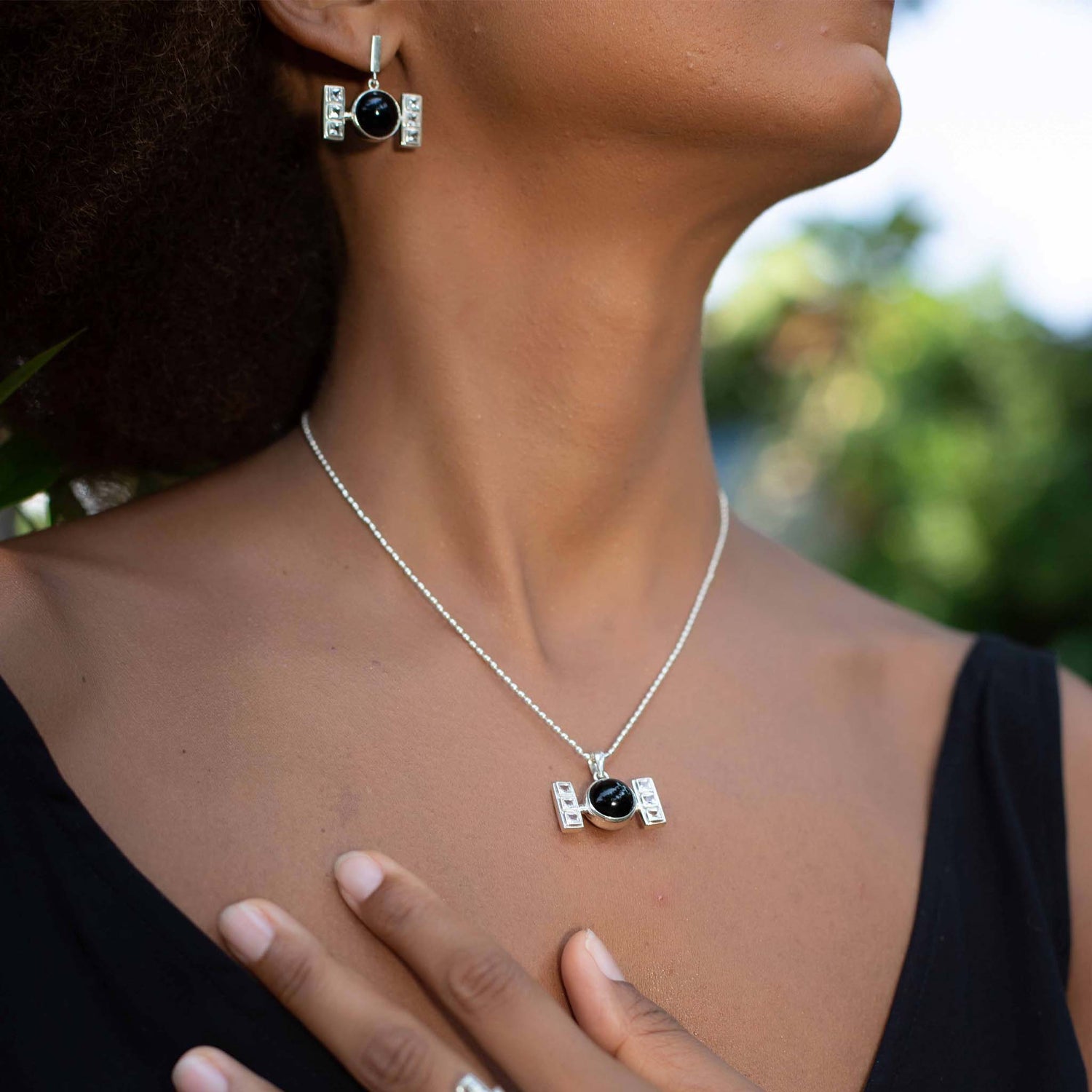 A person wears matching Kaora Sandara Jewelry featuring sterling silver and black agate accents, including earrings and the Health Formula Pendant - Sterling Silver with Black Agate & Crystal. The Health Formula pendant and earrings have a symmetrical design with small rectangular silver details flanking the black stone. The person wears a black top.