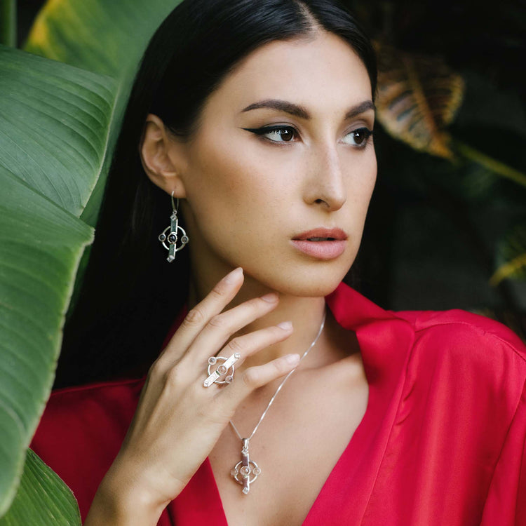 A woman with long dark hair is wearing Kaora Sandara Jewelry's Inner Clarity Earhooks—Sterling Silver 925 with exquisite Crystal Gemstones. Adorned in a red blouse and posing near large green leaves, she creates a natural and elegant backdrop, further complemented by her necklace and ring with Transparent Rhinestone Crystals.