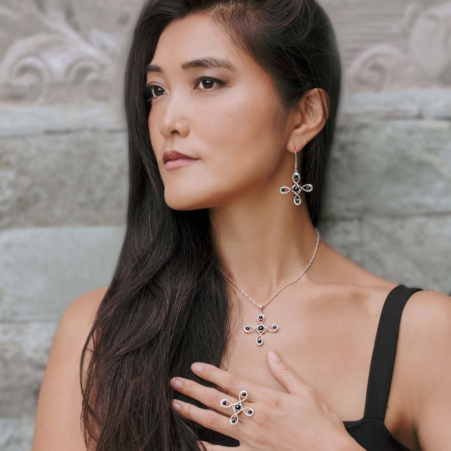 A woman with long, dark hair wears the Programs Clearing Earhooks – Sterling Silver with Black Agate by Kaora Sandara Jewelry, along with a matching Black Agate Sterling Silver 925 necklace and a ring. She is dressed in a sleeveless black top and poses against a stone wall backdrop, with her right hand touching her necklace and wearing a thoughtful expression.