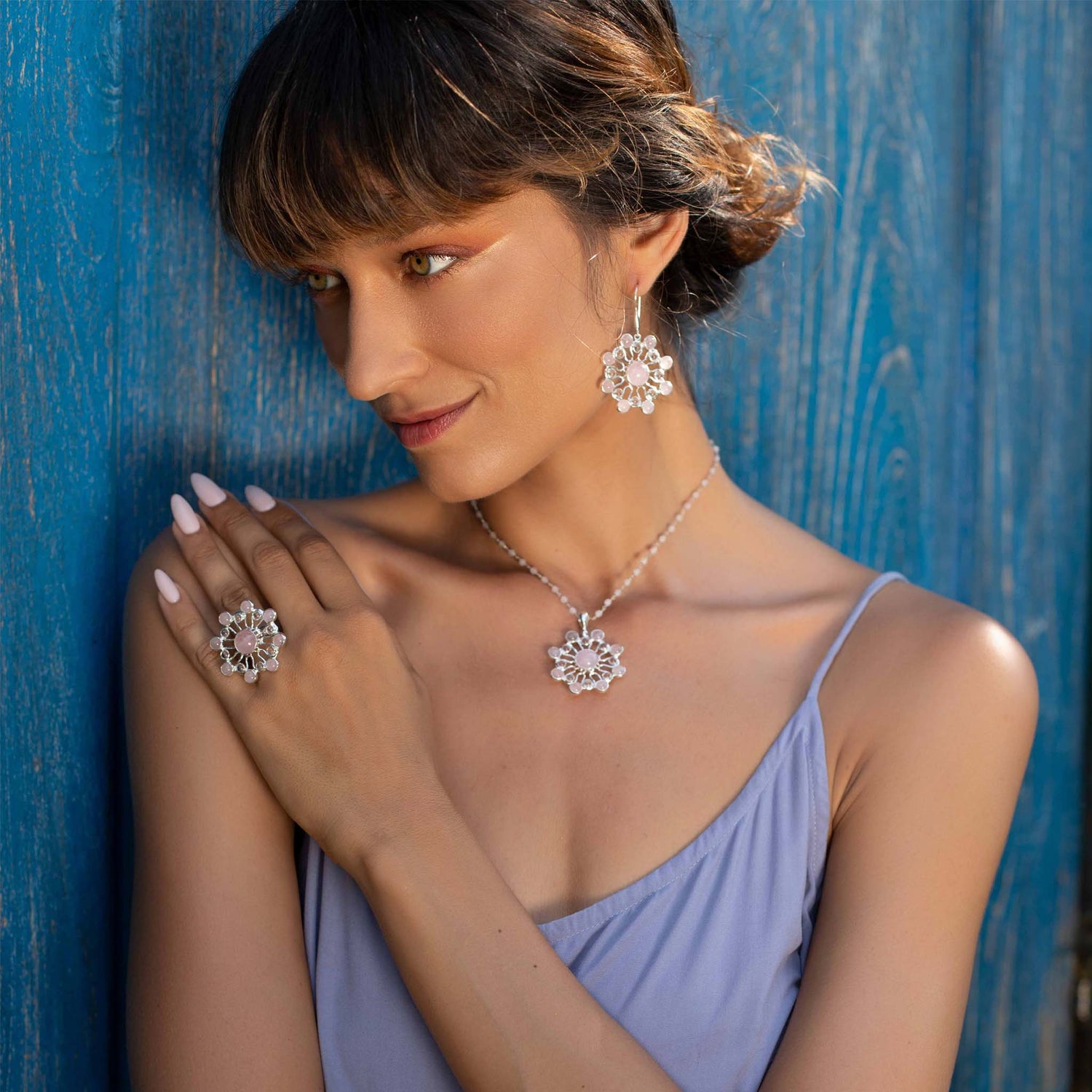 A woman with short brown hair and bangs leans against a blue wall. She is wearing a light blue spaghetti-strap dress, floral earrings, a matching necklace, and the Source of Love Ring - Sterling Silver with Crystal & Rose Quartz by Kaora Sandara Jewelry. She gently touches her shoulder and gazes downward, showcasing the jewelry.