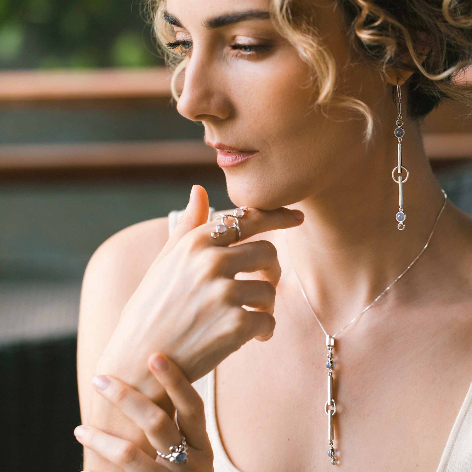 A woman with curly hair is shown thoughtfully looking down. She wears matching silver jewelry from Kaora Sandara Jewelry, including a State Pendant - Sterling Silver with Crystal & Chalcedony necklace, earrings, and rings, all adorned with small Chalcedony Sterling Silver accents. The background is blurred, featuring hints of greenery and a wooden structure.